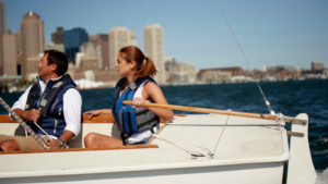 Two people wearing life vests are sailing a small boat on a body of water with a city skyline in the background under a clear blue sky.