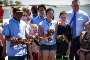 A diverse group of smiling people, including children and adults, stand outdoors by the water on a sunny day. Three people in blue shirts proudly hold large lobsters, while others gather around them.