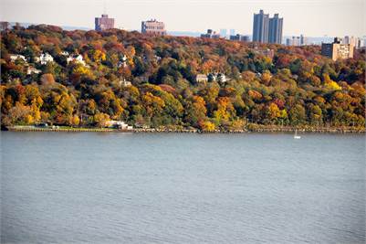 A wide river flows in the foreground with a tree-covered hillside behind, displaying autumn foliage in shades of green, yellow, and orange. Several buildings rise above the trees in the background.