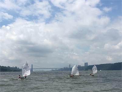 Three small sailboats with white sails are sailing on a wide river, with a distant bridge and city skyline visible under a partly cloudy sky. The water appears slightly choppy.