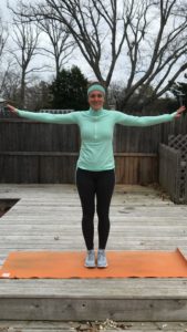A woman in athletic wear stands on an orange mat outdoors with arms outstretched to the sides, smiling, against a backdrop of a wooden deck, fence, and leafless trees.