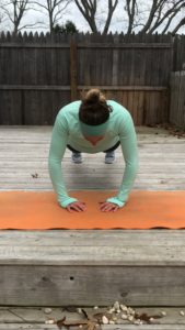 A woman in a light green long-sleeve top and headband is performing a plank with her hands close together, hovering above an orange yoga mat on a wooden deck outdoors.