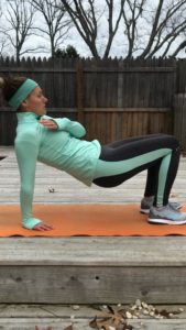 A woman wearing athletic clothes performs a tricep dip exercise on an orange mat outdoors, with one hand on her chest and legs bent. Bare trees and a wooden fence are visible in the background.