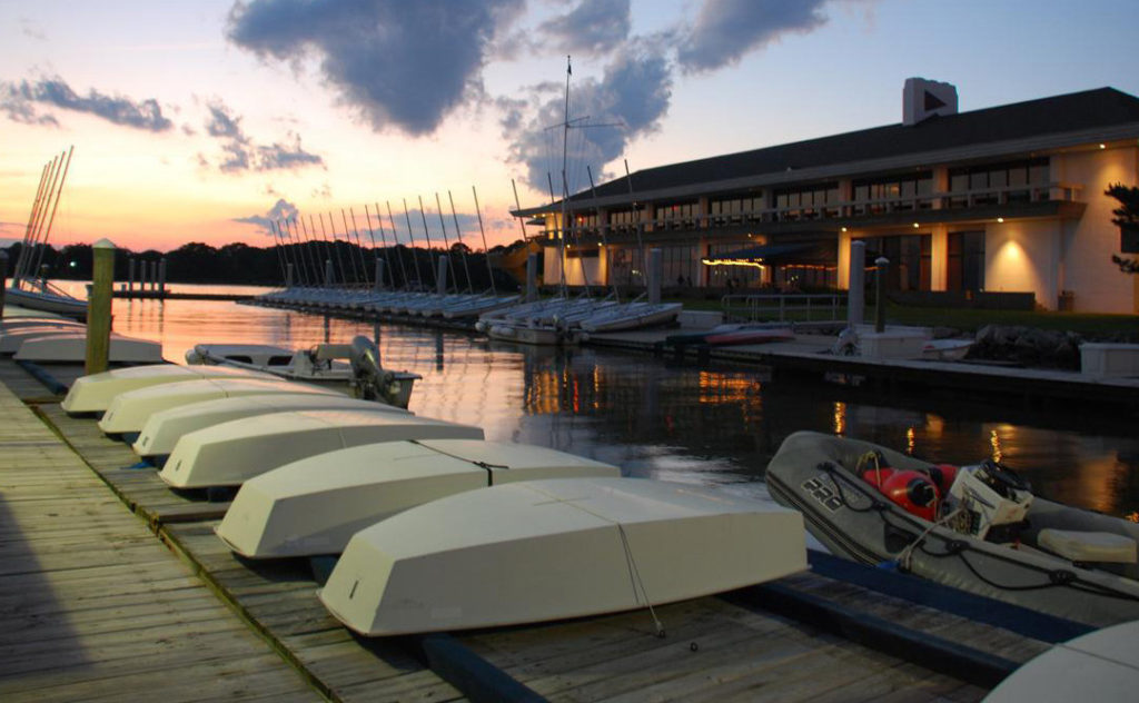 Rowboats are lined up on a wooden dock beside a calm marina at sunset. A large building with many windows overlooks the water, and sailboats are moored in the background. The sky is lit with orange and purple hues.