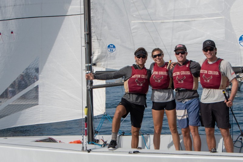 Four young adults wearing sunglasses and Boston College life vests stand smiling on a sailboat, with white sails and calm blue water in the background on a sunny day.