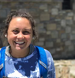 A woman with brown hair smiles at the camera, wearing a blue tie-dye shirt and a bright blue backpack. She stands outdoors in front of a stone wall.