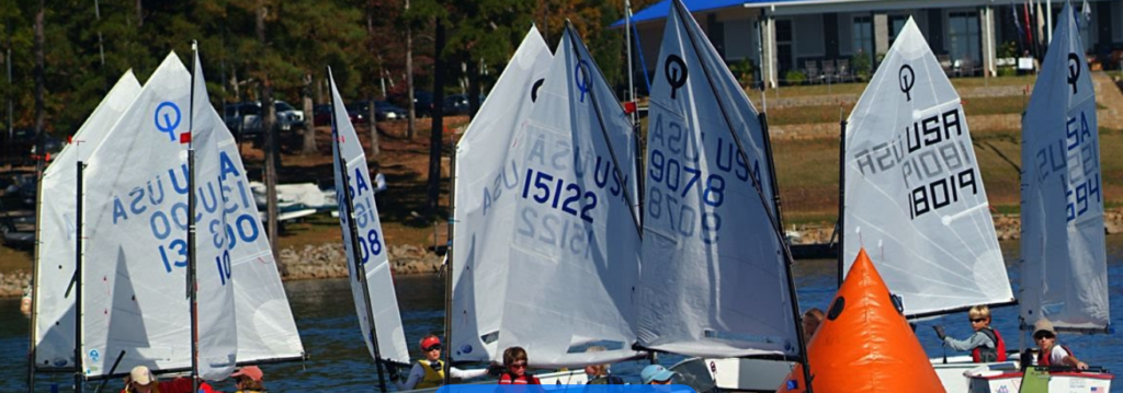 Several small sailboats with white sails, marked with blue and black numbers and "USA," gather near an orange buoy on a lake. Trees and a building are visible on the shore in the background.