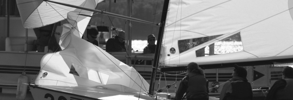 A black and white photo shows several people on and around a sailboat with a crumpled sail, positioned near another boat on the water. The scene appears calm, with sunlight reflecting off the water in the background.