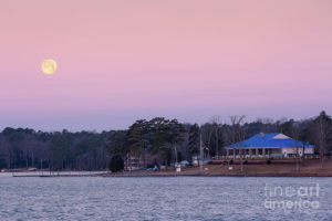 A full moon rises over a calm lake at sunset, with a building featuring a blue roof on the shoreline, surrounded by trees and a pastel-colored sky in the background.