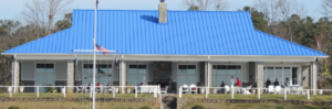 A large building with a bright blue metal roof, wide porch, and white columns. Several people are seated outside on white chairs. An American flag is visible on a pole in front of the building. Trees are in the background.