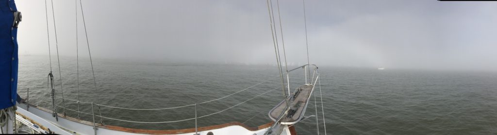 A panoramic view from the deck of a sailboat on calm water, with thick fog obscuring most of the distant horizon and only faint outlines of other boats visible in the mist.