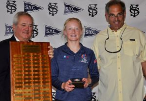 A young woman stands between two men, smiling and holding a small box. One man holds a large wooden plaque with engraved nameplates. They are in front of a backdrop with repeating "S.Y.C." logos and sailing flags.