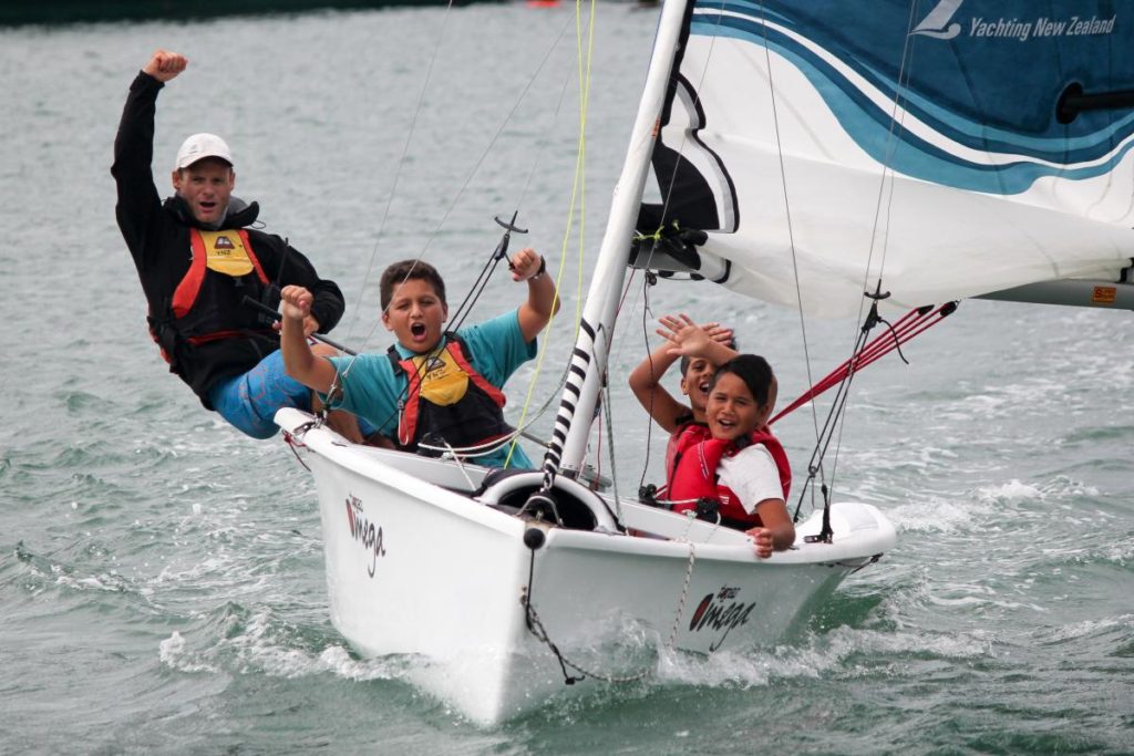 Three children in life jackets cheer enthusiastically while sailing a small white boat on the water, with one raising a fist in excitement and the others smiling and waving. The boat has “Vega” written on it.