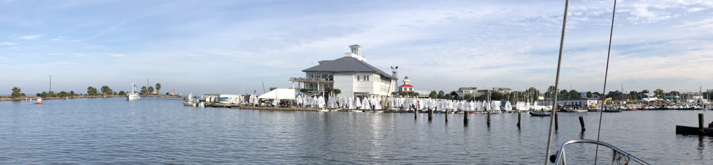 A wide view of a marina with calm water, a large white building, many docked sailboats with folded sails, and a pale blue sky above. Some people and distant buildings are visible onshore.