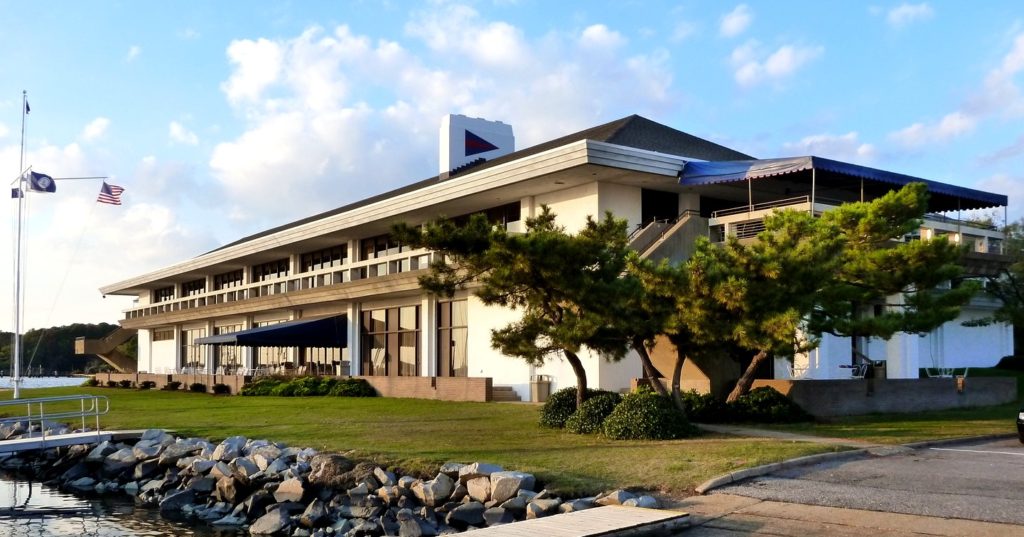 A yacht club building with large windows sits beside the water, surrounded by greenery. An American flag flies on a pole, and a rocky shoreline with a dock appears in the foreground. The sky is partly cloudy.