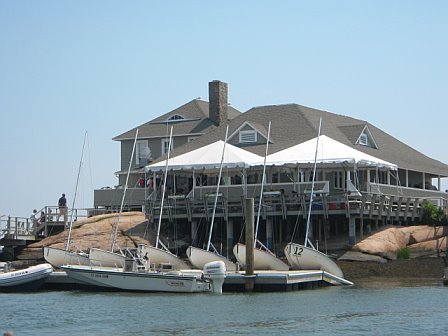 A waterfront building with a large deck and white canopy, overlooking sailboats and a small motorboat docked along the rocky shoreline on a sunny day.