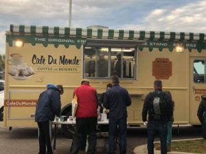A group of people stand in line at a yellow Café Du Monde food truck with a green and white striped awning, ordering coffee and beignets on a cloudy day.