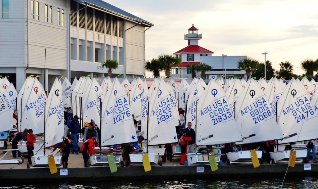 A group of young sailors prepares Optimist dinghies with numbered sails at a marina dock, with a waterfront building and a red-roofed lighthouse in the background. Palm trees line the area.