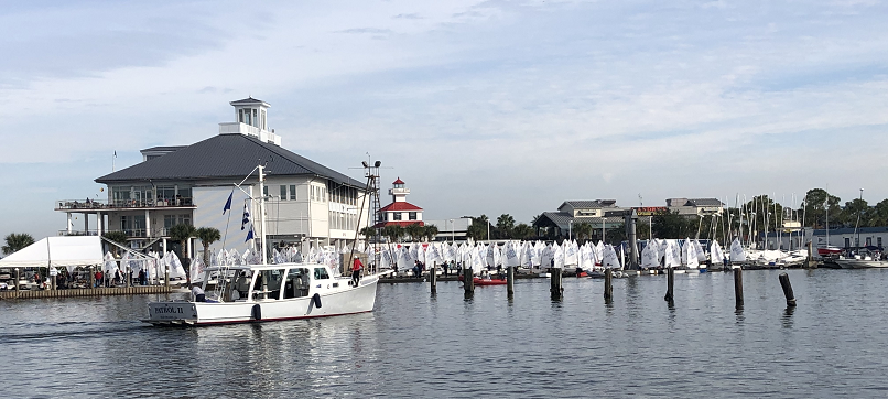 A white boat with blue flags sails on a marina, with a large white building, a red-roofed lighthouse, and many small sailboats docked in the background under a partly cloudy sky.