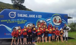 A group of children and two adults wearing life jackets stand smiling in front of a blue trailer with "SAILING...HAVE A GO!" written on it, promoting sailing for kids nationwide.
