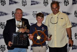 Three people stand in front of an S.Y.C. backdrop, smiling. The person on the left holds a large trophy, the person in the middle holds a wooden ship's wheel award, and the person on the right wears glasses and a yellow shirt.