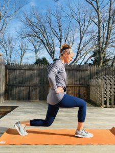 A woman in athletic wear performs a lunge exercise on an orange mat outdoors, with a wooden fence and leafless trees in the background under a clear blue sky.
