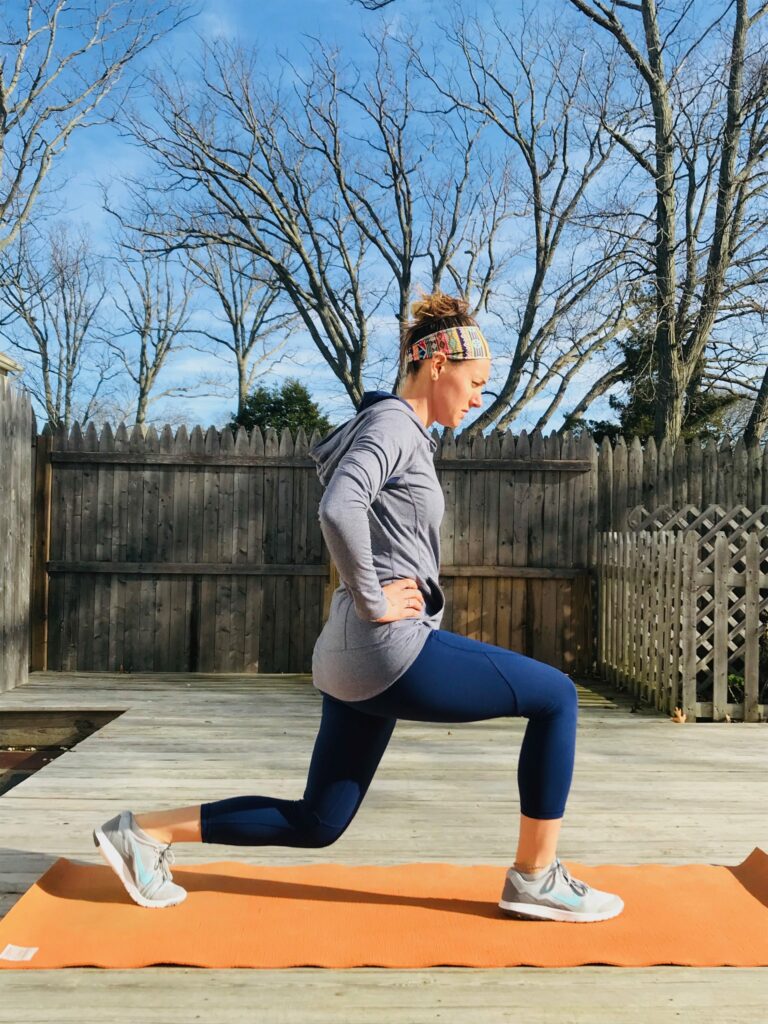A woman in athletic wear performs a lunge exercise on an orange mat outdoors, with a wooden fence and leafless trees in the background under a clear blue sky.