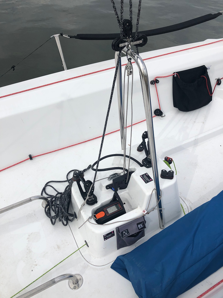 A close-up of a sailboat cockpit shows ropes, control panels, and metal railings. A blue cover lies to the side, and a black storage pouch is attached to the white interior wall of the boat. Water is visible in the background.