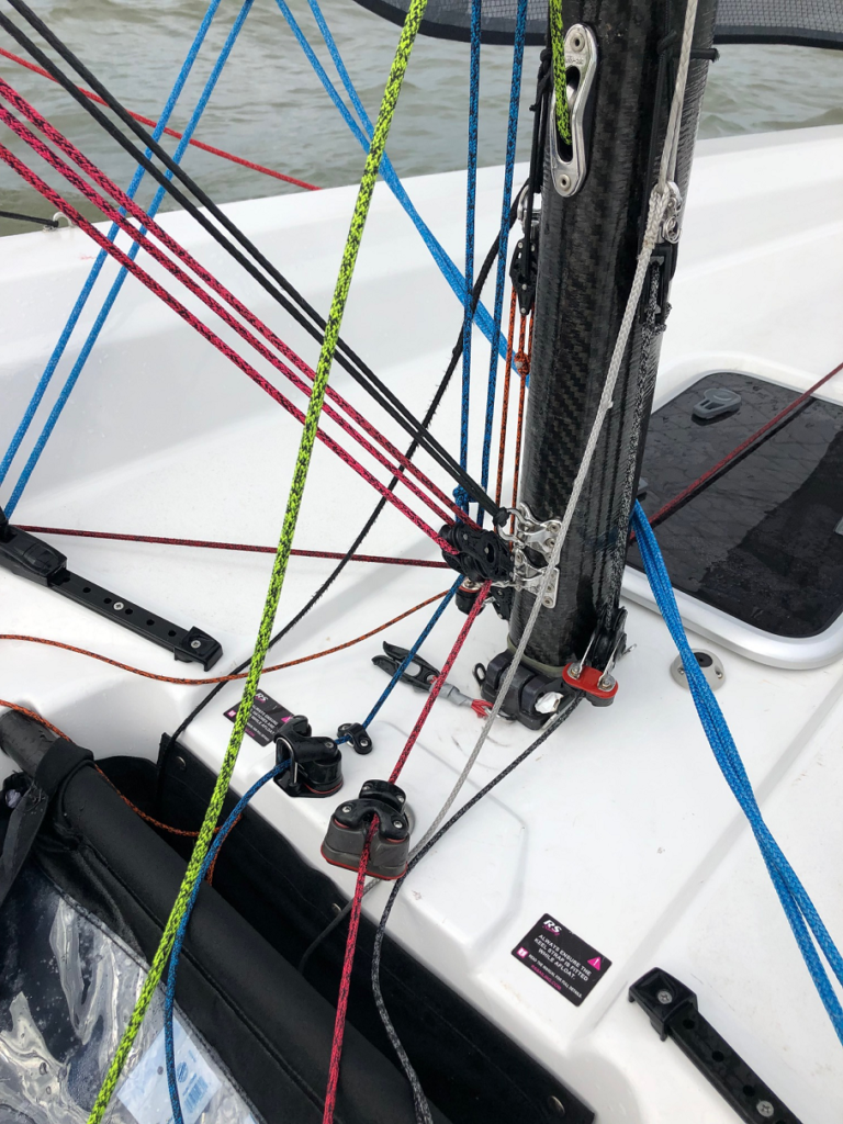 Colorful ropes and rigging are attached to the mast on the deck of a white sailboat, showing various lines and pulleys, with water visible in the background.