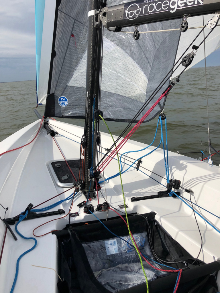 View from inside a small sailboat showing colorful ropes, rigging, and part of the sail on a calm body of water under a cloudy sky. The "racegeek" logo is visible on the sail.