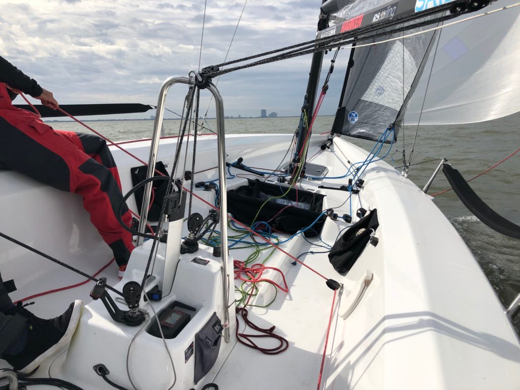 Close-up view of the cockpit of a sailboat in motion, showing ropes, navigation equipment, and a person steering. The sea and a distant city skyline are visible under a cloudy sky.