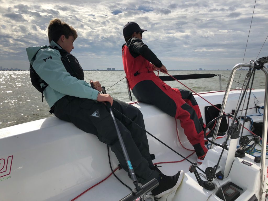 Two people in sailing gear lean out over the side of a sailboat, holding ropes and steering equipment as they navigate on a cloudy day, with calm water and a distant shoreline in the background.