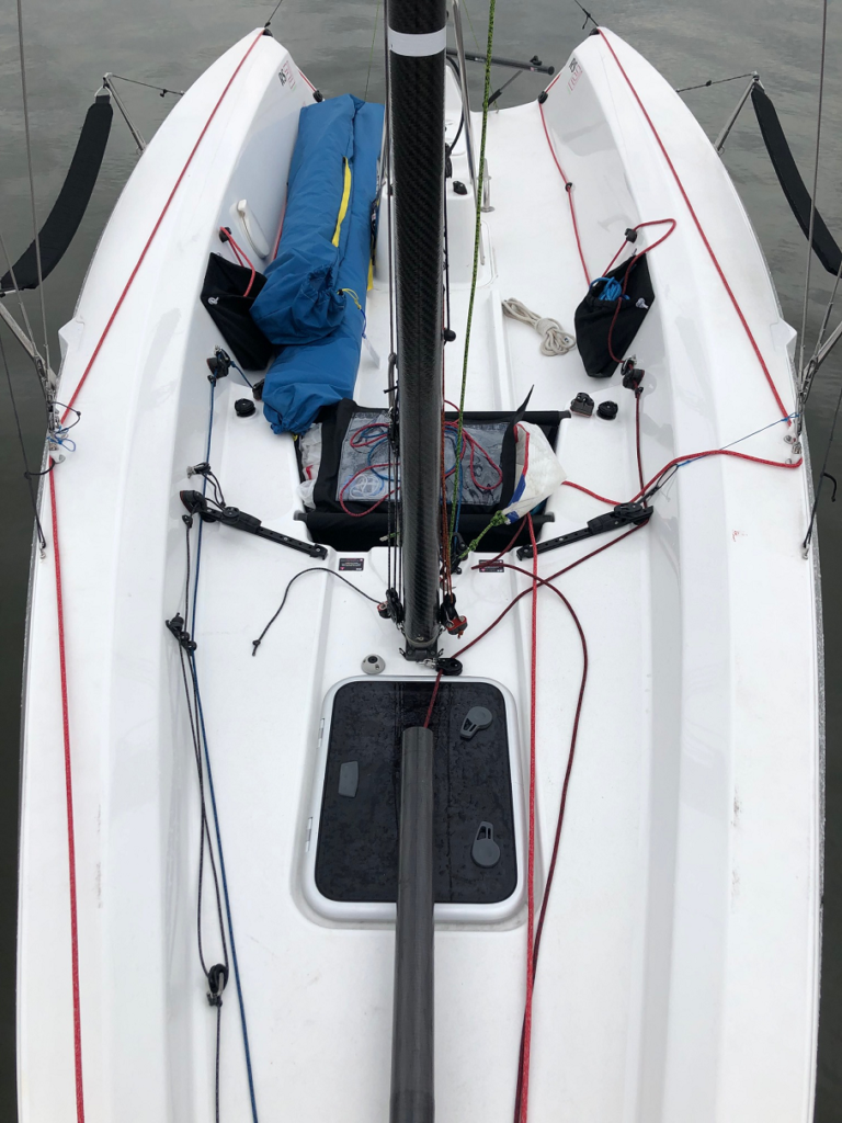 View from above of a white sailboat’s cockpit with various lines, ropes, and gear neatly arranged. A blue sail bag, winches, and a black mast are visible, along with a small hatch and cleats on the deck.