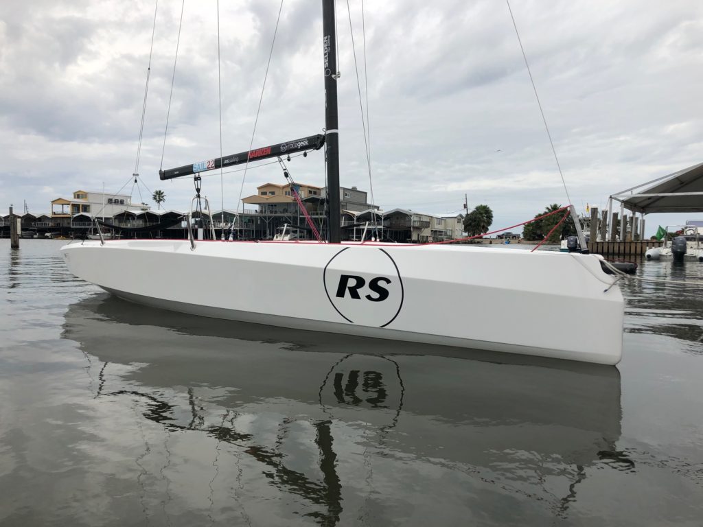 A sleek white sailboat with "RS" in a black circle on its hull floats on calm water near a dock, with houses and a cloudy sky in the background.