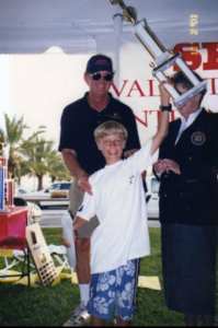 A smiling boy in a white shirt and blue shorts raises a large trophy above his head. An older man in sunglasses stands behind him, and another person in a blazer stands nearby. They are outdoors with palm trees in the background.