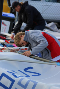 A young person in a gray and red jacket is assembling or inspecting a sailboat, closely examining a sail with blue letters. Another individual works in the background near a fence and parked vehicles.