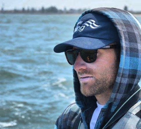 Man wearing a blue plaid hoodie, navy cap, and sunglasses sits by the water on a boat, with blurred trees in the distant background. He has a beard and appears to have sunblock on his lips.