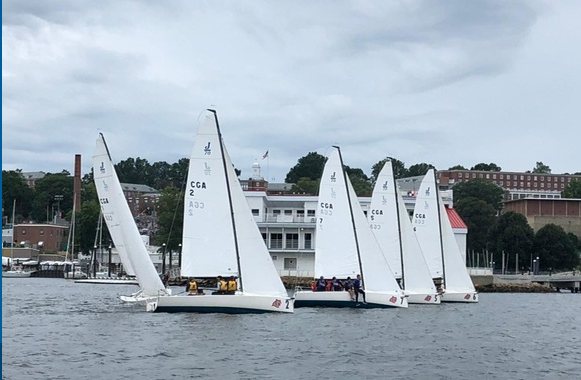 Several white sailboats with numbers on their sails line up on the water near a marina, with a building, trees, and cloudy sky in the background. People can be seen on the boats, preparing to sail.