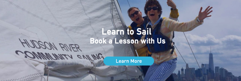 Two people in life jackets smile and pose on a sailboat with 'Hudson River Community Sailing' on the sail. The New York City skyline is visible in the background. Text reads: Learn to Sail. Book a Lesson with Us.
