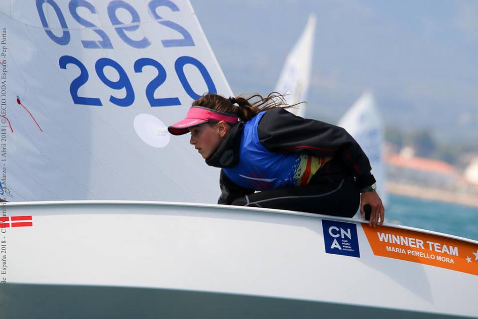 A young woman wearing a visor and blue sailing vest leans forward on a sailboat with the number 2920. The boat displays a sign reading "WINNER TEAM Maria Perello Mora." The background shows water and blurred land.