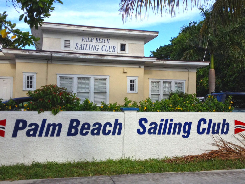 A cream-colored building with white trim and palm trees in front has a sign reading "Palm Beach Sailing Club" on both the building and a wall at the entrance. The scene is bright and tropical.