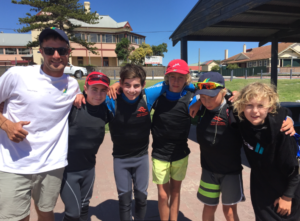 A group of five boys in wetsuits and a man in sunglasses and a white shirt pose together outdoors, smiling under a sunny sky with buildings and trees in the background.