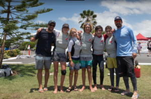 A group of seven people, some wearing life vests and sailing attire, stand barefoot on grass smiling for a photo outdoors, with palm trees and a blue sky in the background.