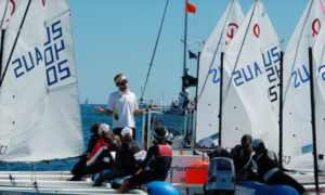 A group of children in life jackets sits in small sailboats on the water, gathered around an adult instructor standing on a motorboat, with white sails raised and blue sky in the background.