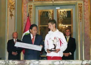 A young athlete in a white tracksuit holds a trophy while standing next to a man in a suit presenting a large check. Two other men stand in the background beside a Peruvian flag in an ornate room.