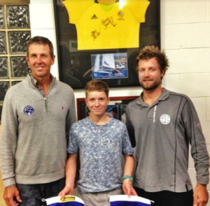 Three people stand indoors smiling at the camera. Above them, a yellow signed sports jersey is displayed on the wall, along with framed photos. The person in the center holds two ribbon-like items.
