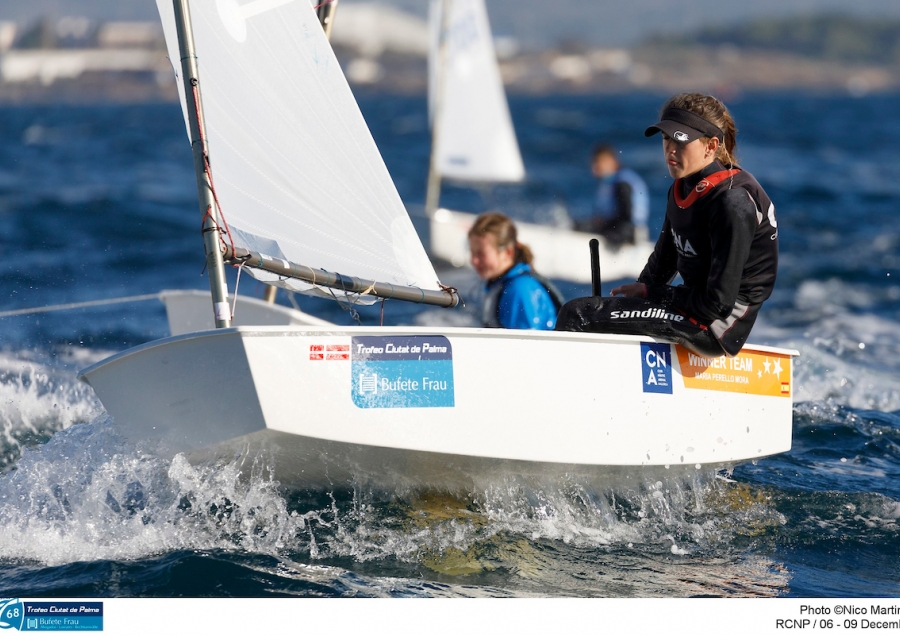 A young sailor wearing sunglasses and a life jacket steers a small white sailboat on choppy blue water, with other sailors and boats visible in the background.
