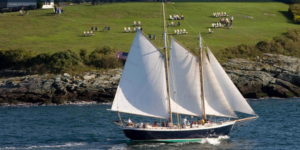 A large sailboat with white sails glides across the water near a rocky shore, with a grassy hill and people sitting on chairs in the background. An American flag flies from the boat’s stern.