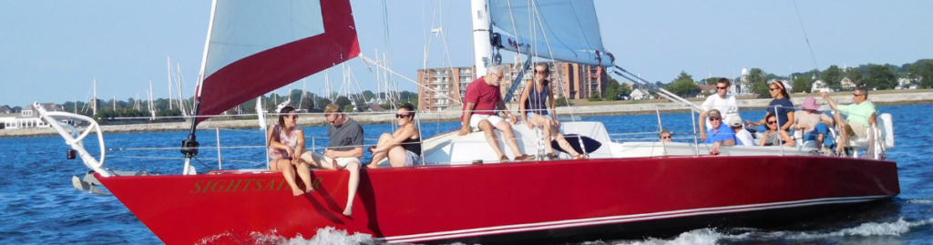 A group of people relax on the deck of a red sailboat named "Sightsail" as it sails on calm blue water near a marina, with shoreline buildings and trees visible in the background.