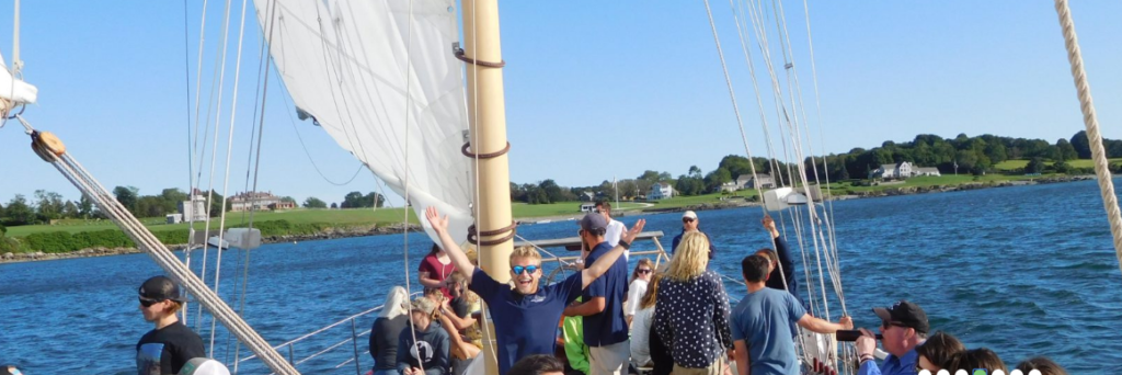 A group of people enjoy a sunny day on a sailboat, some standing and others sitting, with blue water and a green shoreline in the background. The sail is up and the sky is clear.